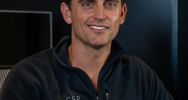 A man, smiling, sits with his hands folded