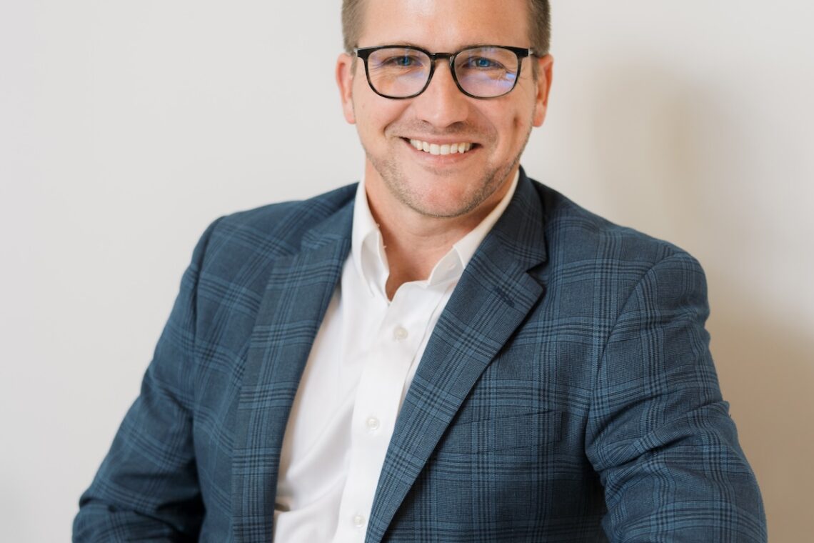 A professional headshot of a smiling man with short brown hair and glasses, wearing a blue plaid blazer, white button-down shirt, and khaki pants while seated at a white table.