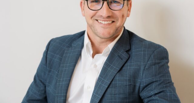 A professional headshot of a smiling man with short brown hair and glasses, wearing a blue plaid blazer, white button-down shirt, and khaki pants while seated at a white table.