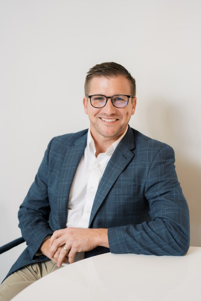 A professional headshot of a smiling man with short brown hair and glasses, wearing a blue plaid blazer, white button-down shirt, and khaki pants while seated at a white table.