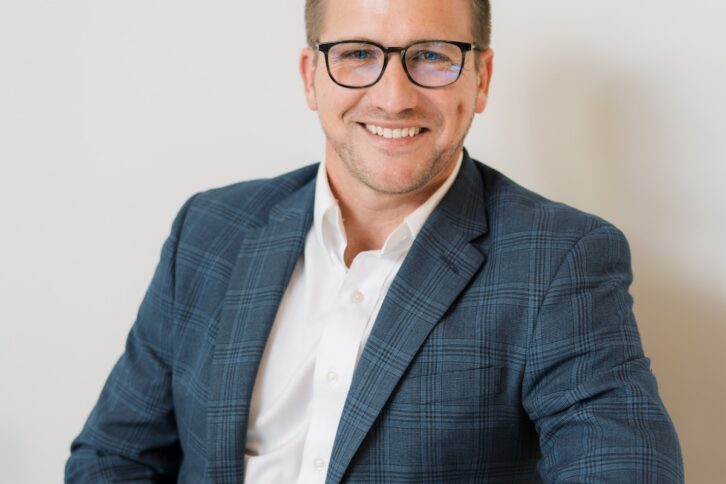 A professional headshot of a smiling man with short brown hair and glasses, wearing a blue plaid blazer, white button-down shirt, and khaki pants while seated at a white table.