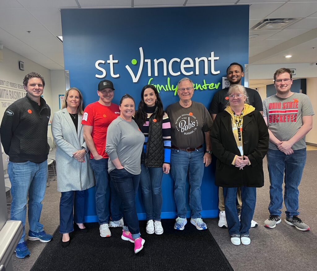 A group of nine smiling individuals poses together indoors in front of a blue wall featuring the "St. Vincent Family Center" logo.