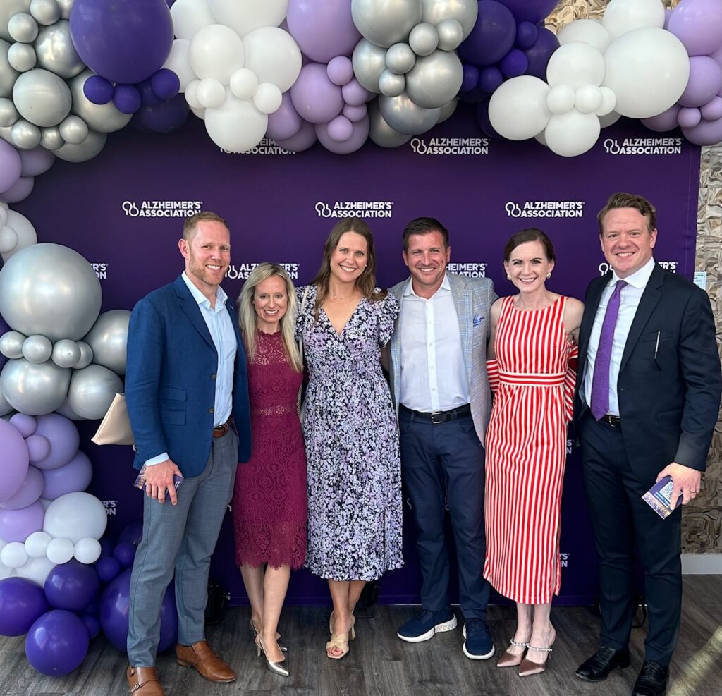 Six people dressed in nice attire smiling for a group photo in front of a purple Alzheimer's Association backdrop framed by purple, white, and silver balloons.