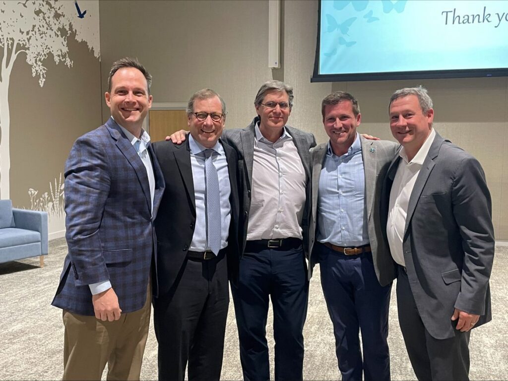Five men in business-casual and formal suits smile together in an auditorium setting, with a projector screen reading "Thank you" visible in the background.